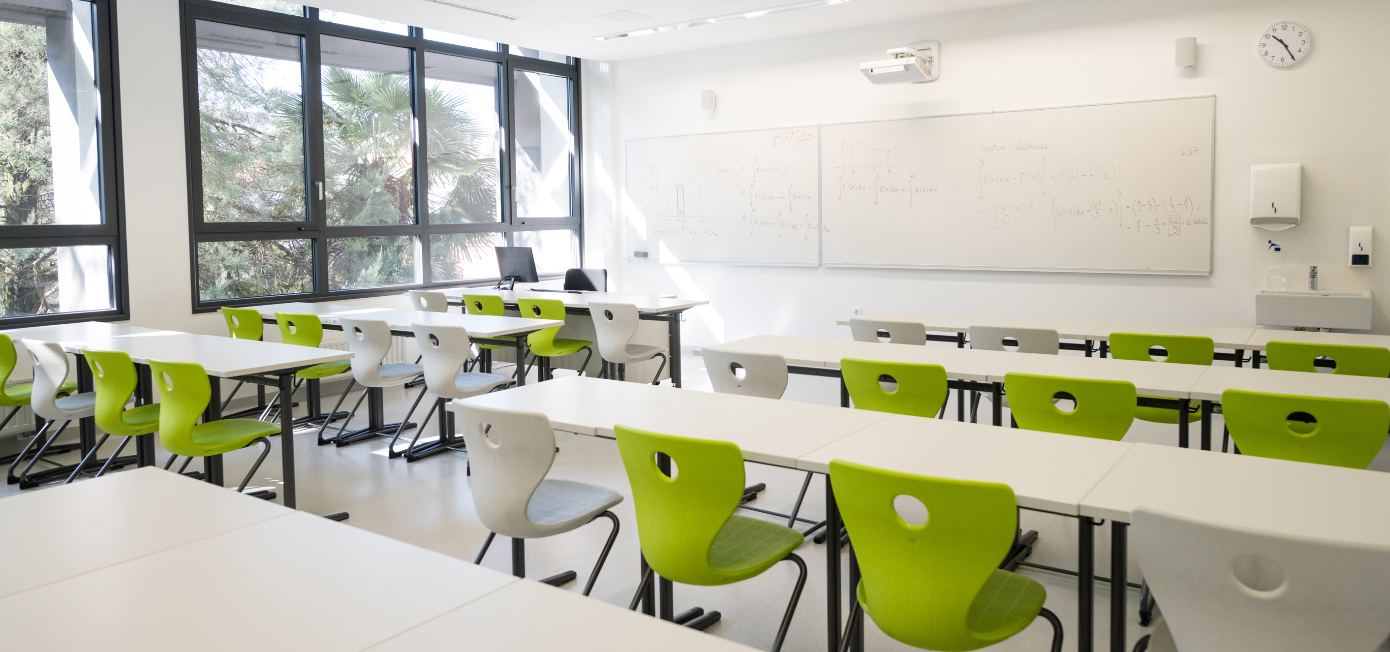 A contemporary classroom, mostly white with some green chairs, some white, and a set of windows looking out over trees.